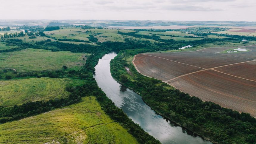 Áreas agrícolas às margens de um rio, sob céu parcialmente nublado, em paisagem rural de São Paulo.