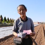 Produtor rural idoso segura um documento e uma pá em uma área agrícola ensolarada.