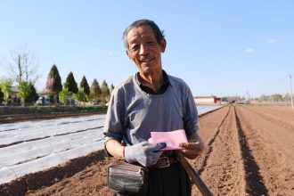 Produtor rural idoso segura um documento e uma pá em uma área agrícola ensolarada.